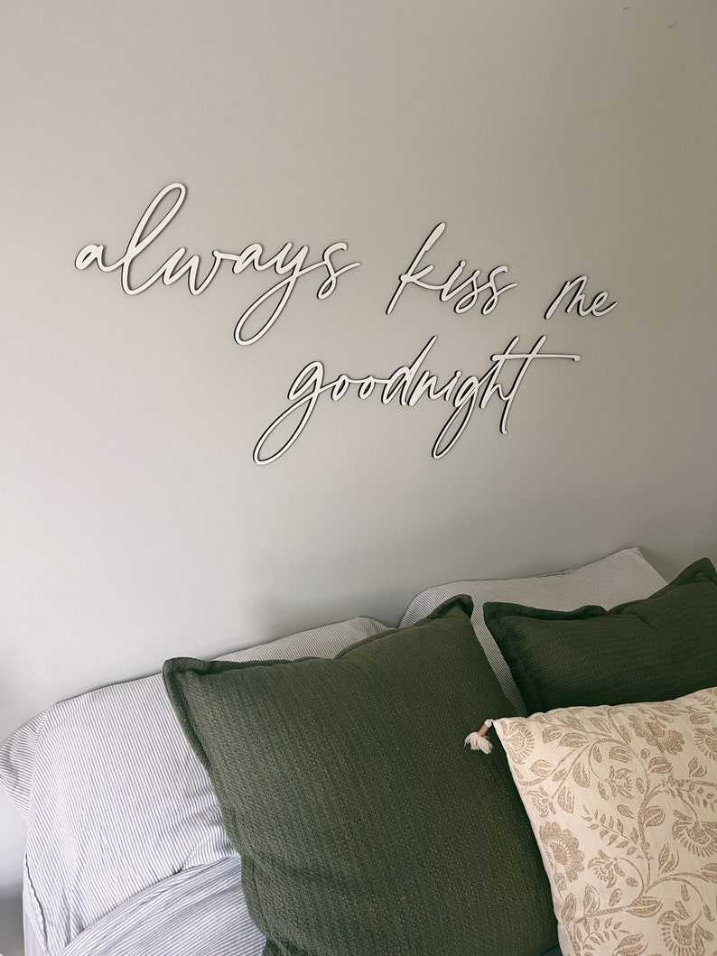 May include: A bedroom with a white wall and a bed with green and white patterned pillows. The wall has a white wooden sign that reads "always kiss me goodnight".