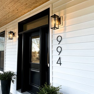 A large, black solid aluminum house number in a thin, modern, and trendy font is mounted on white farmhouse-style siding, offering a striking contrast and contemporary elegance against the classic backdrop.