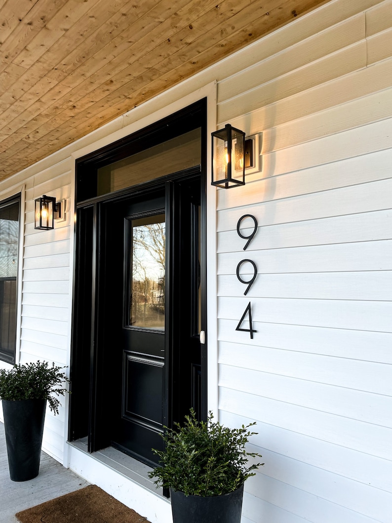 A large, black solid aluminum house number in a thin, modern, and trendy font is mounted on white farmhouse-style siding, offering a striking contrast and contemporary elegance against the classic backdrop.