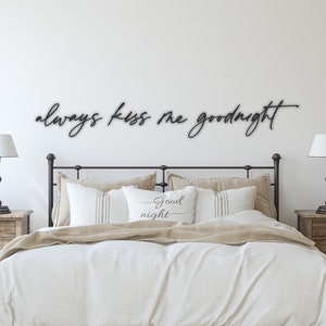 May include: A bedroom with a white wall and a black metal sign that reads "always kiss me goodnight". The sign is above a bed with white bedding and a pillow that reads "good night". There are two wooden bedside tables with lamps on either side of the bed.