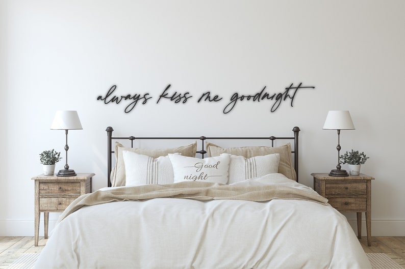 May include: A bedroom with a white wall and a black metal sign that reads "always kiss me goodnight". The sign is above a bed with white bedding and a pillow that reads "good night". There are two wooden bedside tables with lamps on either side of the bed.