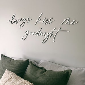 May include: White wooden sign with the text "always kiss me goodnight" on a white wall above a bed with green and white pillows.