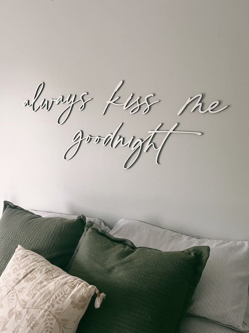 May include: White wooden sign with the text "always kiss me goodnight" on a white wall above a bed with green and white pillows.