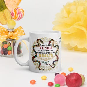 Puede incluir: Taza de cerámica blanca con asa, con un diseño de dos serpientes, texto que dice "MAY ALL THE VENOM SPOKEN ABOUT ME BE TRANSMUTED INTO WEALTH" y la frase "SO BE IT".