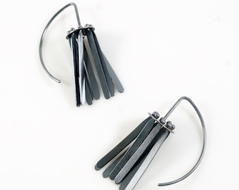 May include: A pair of silver-toned earrings. Each earring features a curved ear wire and a cluster of small, dark gray rectangular bars. The bars are arranged in a fringe-like design, creating a modern and minimalist aesthetic.