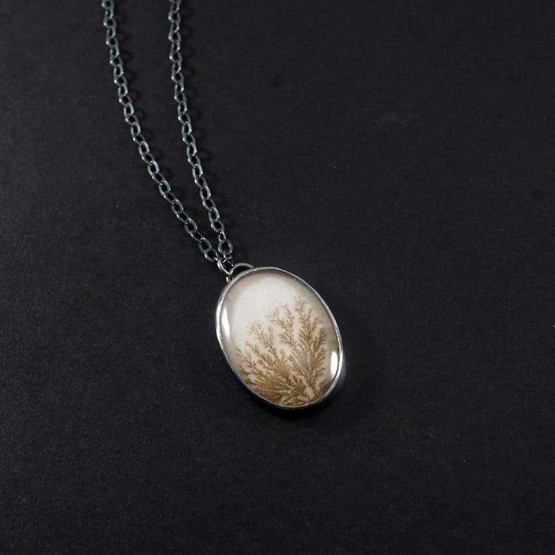 May include: Oval pendant necklace with a silver chain. The pendant features a white background with a brown, tree-like pattern encased in a silver bezel. The necklace is displayed against a black background.