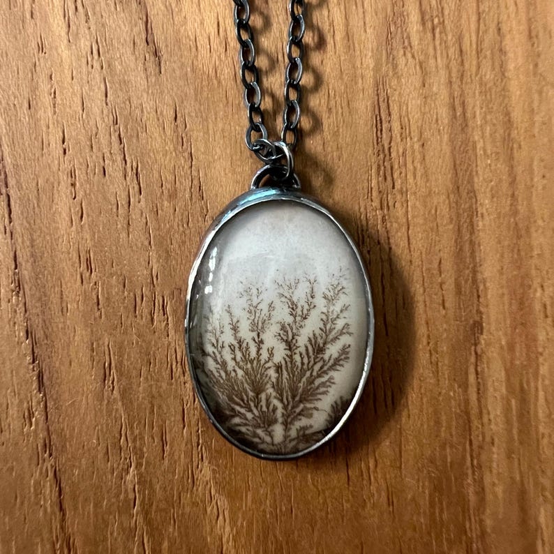 May include: Oval pendant necklace with a silver-tone chain. The pendant features a white background with a brown, fern-like pattern. The necklace is displayed on a wooden surface.