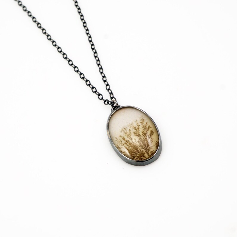 May include: Oval pendant necklace with a silver-tone chain. The pendant features a cream-colored stone with a brown, tree-like pattern. The pendant is framed in a silver-tone metal.