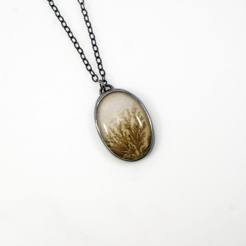 May include: Oval pendant necklace with a silver-tone chain. The pendant features a cream-colored stone with a brown, fern-like pattern. The pendant is encased in a silver-tone metal frame. The necklace is displayed against a white background.