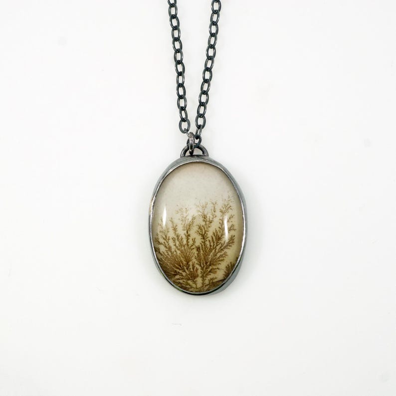 May include: Oval pendant necklace with a silver-tone chain. The pendant features a cream-colored stone with a brown, fern-like pattern. The pendant is encased in a silver-tone metal frame. The necklace is displayed against a white background.