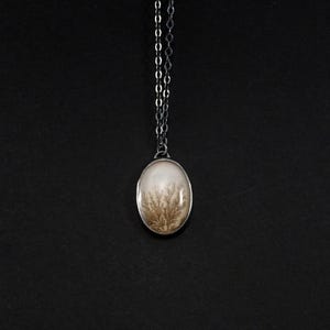 May include: Oval pendant necklace featuring a cream-colored stone with a dendritic pattern in shades of brown. The stone is set in a silver-toned metal frame and hangs from a delicate silver chain. The background is solid black.