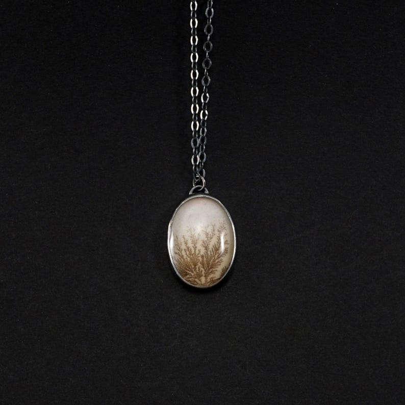 May include: Oval pendant necklace featuring a cream-colored stone with a dendritic pattern in shades of brown. The stone is set in a silver-toned metal frame and hangs from a delicate silver chain. The background is solid black.