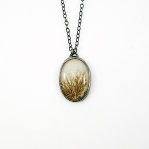 May include: An oval pendant necklace with a silver-colored chain. The pendant features a cream-colored background with a brown, fern-like pattern. The pendant is encased in a silver-colored metal frame.