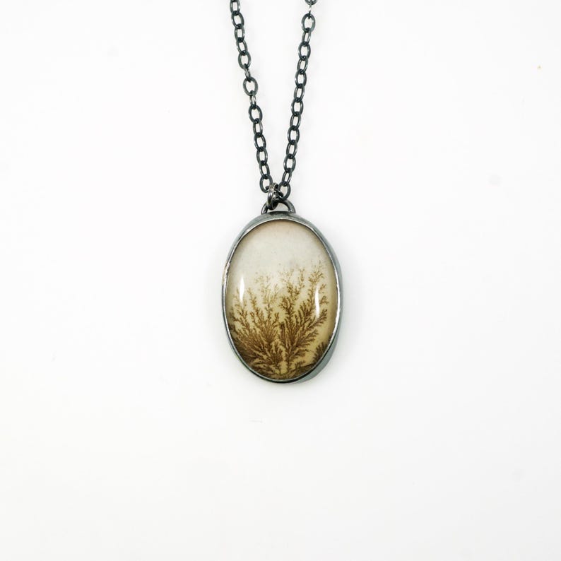 May include: An oval pendant necklace with a silver-colored chain. The pendant features a cream-colored background with a brown, fern-like pattern. The pendant is encased in a silver-colored metal frame.