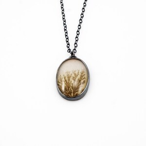 May include: Oval pendant necklace with a natural dendritic agate stone. The stone features brown fern-like inclusions set in a cream-colored background. The pendant is framed in a silver-colored metal and hangs from a dark chain.