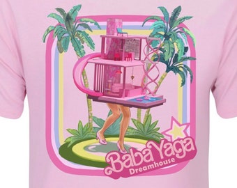 Puede incluir: Camiseta rosa con un gráfico de una casa rosa con piernas de mujer que salen de la parte inferior. La casa está rodeada de palmeras y el texto "Baba Yaga Dreamhouse" está debajo de la casa.