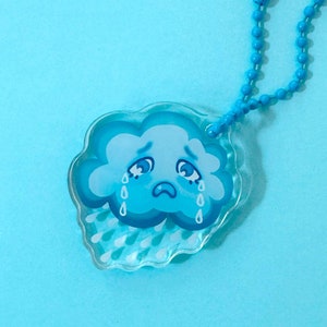 May include: A clear acrylic keychain featuring a cartoon cloud with a sad face and teardrops. The cloud is light blue with darker blue accents. A matching blue beaded chain is attached to the keychain.