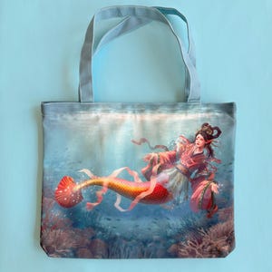 May include: A tote bag with a colorful illustration of a mermaid with a red, orange, and yellow tail. The mermaid is wearing a pink and white dress and has long black hair. The background is a blue ocean with coral reefs and fish.
