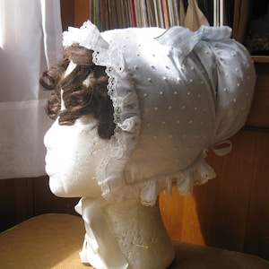 May include: A white lace bonnet with blue and white striped ribbon. The bonnet is made of a soft, white fabric with a dotted pattern.