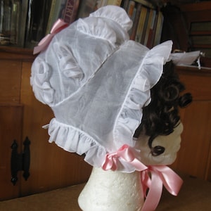 May include: A white lace bonnet with pink ribbon trim. The bonnet is worn on a white mannequin head with brown curly hair.