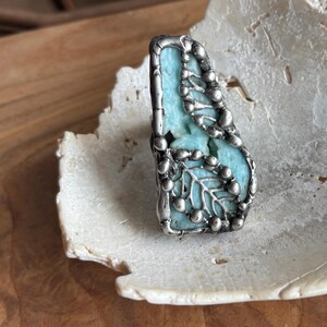 May include: A rectangular pendant with a light blue stone center, framed by a silver metal border. The silver metal features a leaf and berry design on one side. The pendant is set on a white, textured surface.