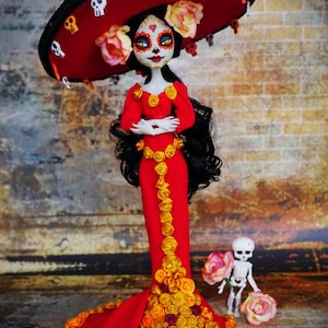 Puede incluir: Muñeca artesanal del Día de Muertos con un vestido rojo adornado con rosas amarillas, un gran sombrero rojo con detalles de calaveras y una pequeña figura de esqueleto. La muñeca tiene una cara pintada con detalles intrincados.
