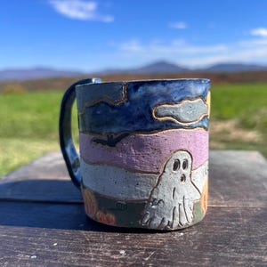 May include: A ceramic mug with a dark blue handle featuring a hand-painted scene of a white ghost, orange pumpkins, and a black bat against a sky with blue clouds and a purple horizon. The mug has a textured surface and a glossy finish.