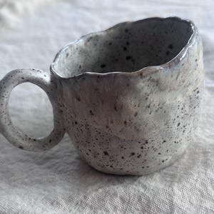 May include: A handmade ceramic mug with a speckled, textured surface. The mug is primarily gray with dark brown speckles and a rounded handle. The rim has a slightly irregular shape, adding to its unique, artisanal appearance.
