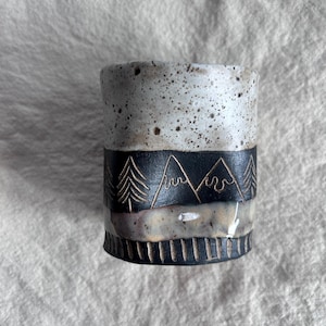 May include: A handmade ceramic tumbler with a speckled white top, a black band featuring etched mountain and tree designs, and a textured base. The cup is cylindrical and has a rustic, earthy aesthetic.