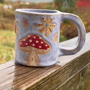 May include: A handmade ceramic mug with a light purple glaze. The mug features a red mushroom design with white spots and gold accents. Gold stars and floral designs are also painted on the mug. The mug is sitting on a wooden surface.