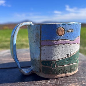 May include: A ceramic mug with a textured, speckled surface featuring a hand-painted landscape design. The scene depicts a blue sky with a brown sun, purple and white rolling hills, and green fields with small trees and grass at the base.
