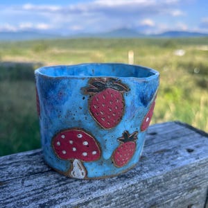 May include: A handmade ceramic cup with a bright blue glazed finish, featuring hand-painted red strawberries with green stems and a red mushroom with white spots. The cup has an uneven, organic rim and a speckled texture throughout the blue surface.