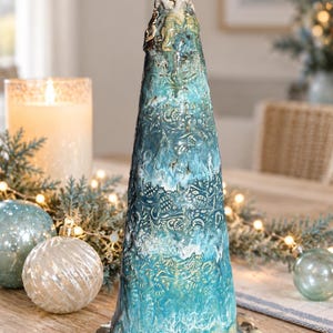 May include: A conical ceramic sculpture featuring a textured surface with embossed starfish and shell motifs. The piece displays a gradient of blue, turquoise, and white glazes, topped with a cluster of sculpted starfish at the apex.