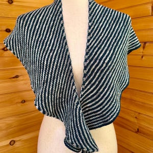 May include: A knitted shawl with a striped pattern in navy blue and white. The shawl is draped over a mannequin, showcasing its design and texture. The garment has short sleeves and a flowing, open front.