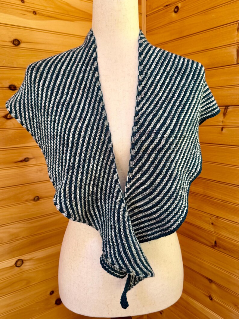 May include: A knitted shawl with a striped pattern in navy blue and white. The shawl is draped over a mannequin, showcasing its design and texture. The garment has short sleeves and a flowing, open front.