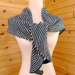 May include: A knitted shawl in teal and off-white stripes, displayed on a mannequin. The shawl is draped to show its shape and texture. The stripes run diagonally across the fabric, creating a visual pattern. The background is a wooden wall.