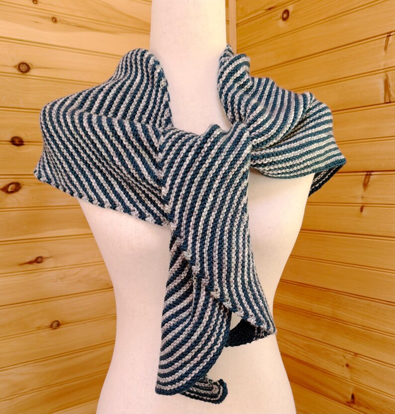 May include: A knitted shawl in teal and off-white stripes, displayed on a mannequin. The shawl is draped to show its shape and texture. The stripes run diagonally across the fabric, creating a visual pattern. The background is a wooden wall.