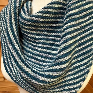 May include: A hand-knitted cowl scarf in teal and white stripes. The soft, textured scarf is wrapped around a mannequin, showcasing the alternating bands of color. The cozy accessory is perfect for adding warmth and style.