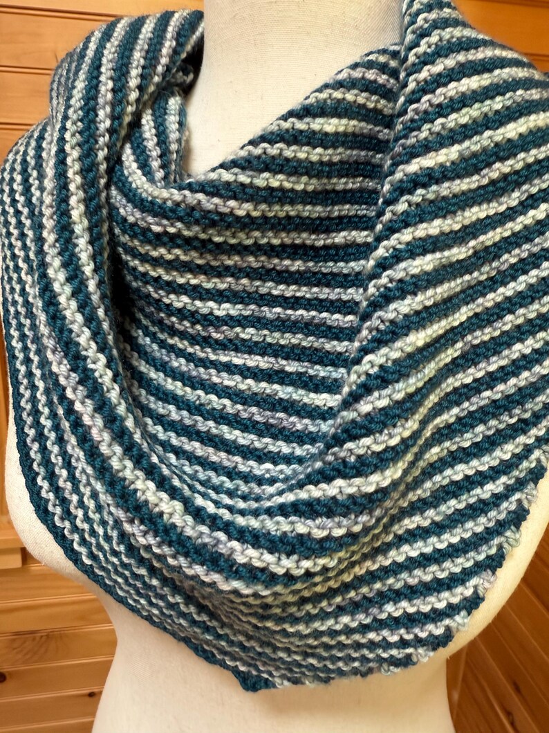May include: A hand-knitted cowl scarf in teal and white stripes. The soft, textured scarf is wrapped around a mannequin, showcasing the alternating bands of color. The cozy accessory is perfect for adding warmth and style.