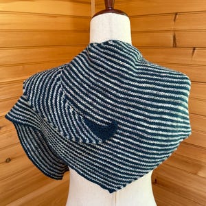 May include: A knitted shawl in navy blue and white stripes is draped over a mannequin. The shawl has a triangular shape and is made of soft yarn. The stripes run horizontally across the fabric, creating a visually appealing pattern. The shawl is a cozy accessory.
