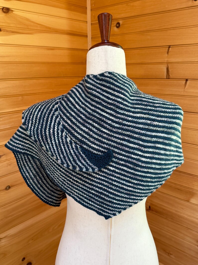 May include: A knitted shawl in navy blue and white stripes is draped over a mannequin. The shawl has a triangular shape and is made of soft yarn. The stripes run horizontally across the fabric, creating a visually appealing pattern. The shawl is a cozy accessory.