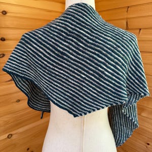 May include: A knitted shawl in shades of teal and white, displayed on a mannequin. The shawl features a striped pattern, with alternating bands of teal and white. The edges are finished with a simple border. The shawl is draped over the mannequin's shoulders.