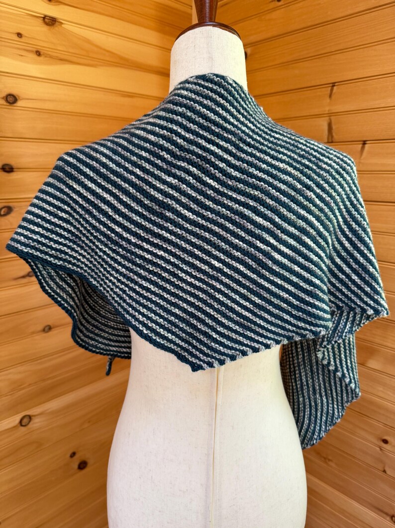 May include: A knitted shawl in shades of teal and white, displayed on a mannequin. The shawl features a striped pattern, with alternating bands of teal and white. The edges are finished with a simple border. The shawl is draped over the mannequin's shoulders.