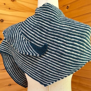 May include: A knitted shawl in shades of teal and white, featuring a striped pattern. The shawl is draped over a mannequin, showcasing its triangular shape and textured design. The fabric appears soft and cozy.