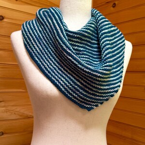 May include: A blue and white striped knitted cowl scarf displayed on a mannequin. The scarf is a triangular shape, with alternating stripes of dark blue and white. The scarf is made of a soft, textured yarn.