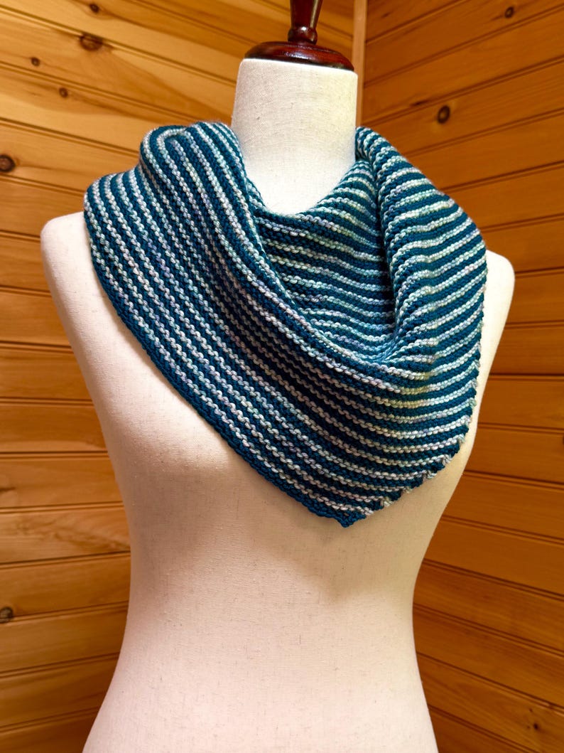 May include: A blue and white striped knitted cowl scarf displayed on a mannequin. The scarf is a triangular shape, with alternating stripes of dark blue and white. The scarf is made of a soft, textured yarn.