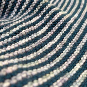 May include: Close-up of a knitted fabric with a striped pattern. The alternating stripes are teal and off-white, creating a textured surface. The knitwear appears soft and flexible, with visible stitch detail.