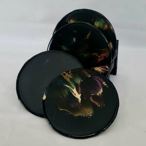 Puede incluir: Conjunto de cuatro posavasos negros con patrones abstractos en espiral en tonos de verde, morado, dorado y blanco.