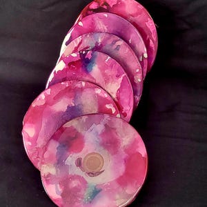 Puede incluir: Un juego de seis posavasos redondos de color rosa y morado con efecto marmoleado. Los posavasos tienen un acabado brillante.