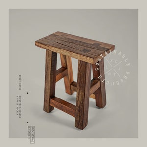 Può includere: Un piccolo sgabello rustico in legno con un sedile rettangolare e quattro robuste gambe. Lo sgabello è realizzato in legno marrone scuro con venature e nodi visibili, che gli conferiscono un aspetto vintage. Il testo "SUSTAINABLE PRODUCTS" è in un cerchio a destra.
